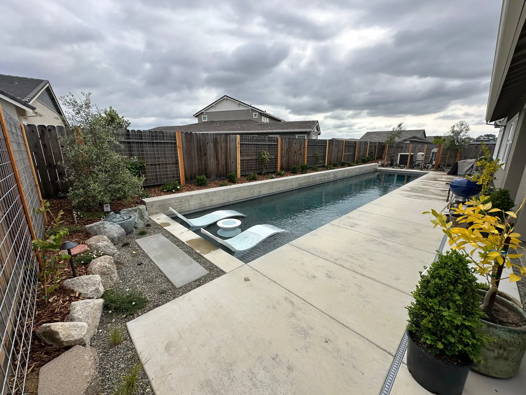 Poolside landscape with concrete patio, boulders, and modern fence